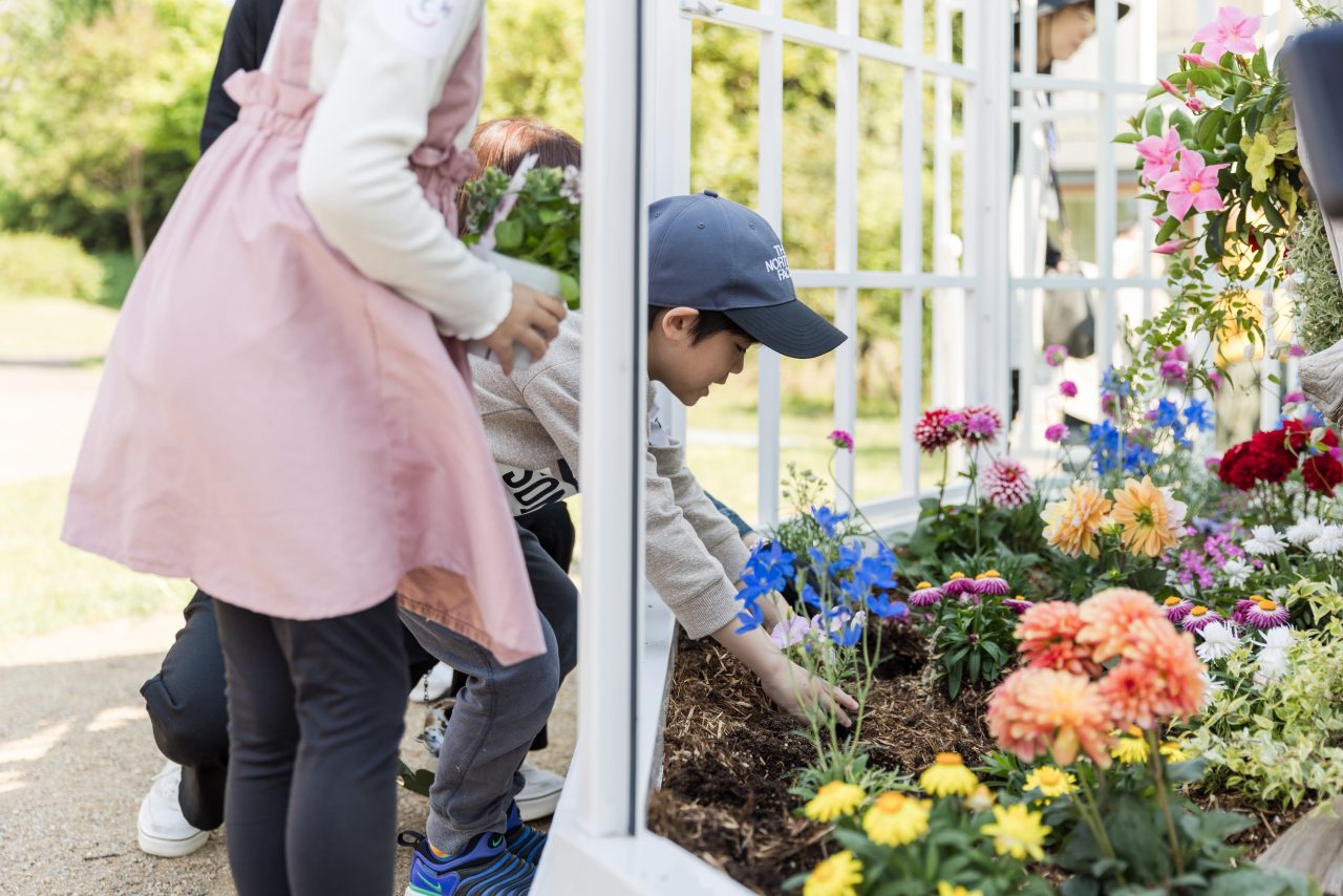 [写真]自分の手で作品の花壇にお花を植える子ども。その周囲には色とりどりの花が植えられています。
