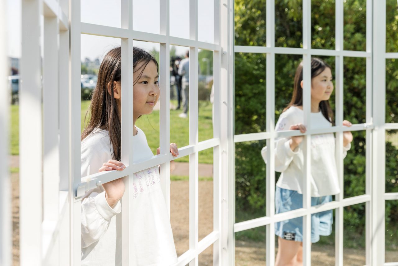 [写真]作品を囲う白い格子から鏡になった作品の中を覗く子ども。鏡にもその子どもの姿が写っています。