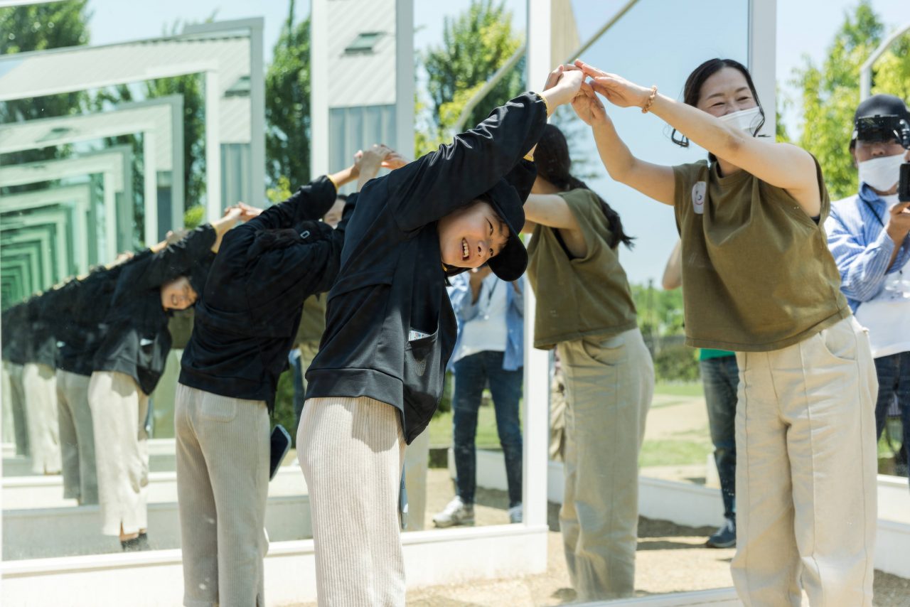 [写真]作品の4つの庭の間の通路で、子どもと大人が手を伸ばして、頭の上でアーチをつくって、合わせ鏡を覗いています。鏡にはどんどんと小さくなっていくふたりの姿が映っています