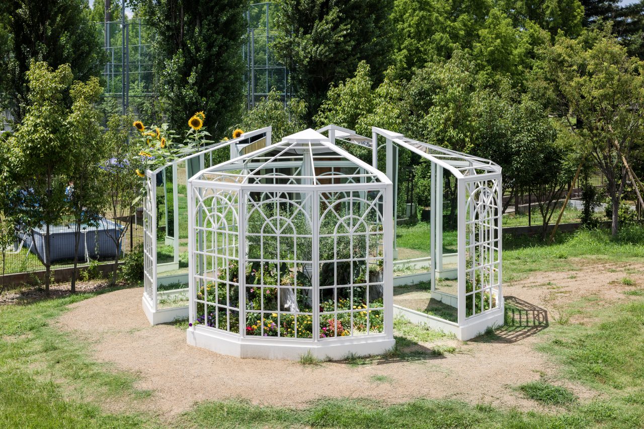 [写真]TSURUMIこどもホスピスの建物側の少し高い位置から作品の外観をうつした写真です。大人の背の高さよりも高い大きな白い鳥かごを4等分したような形をしています。